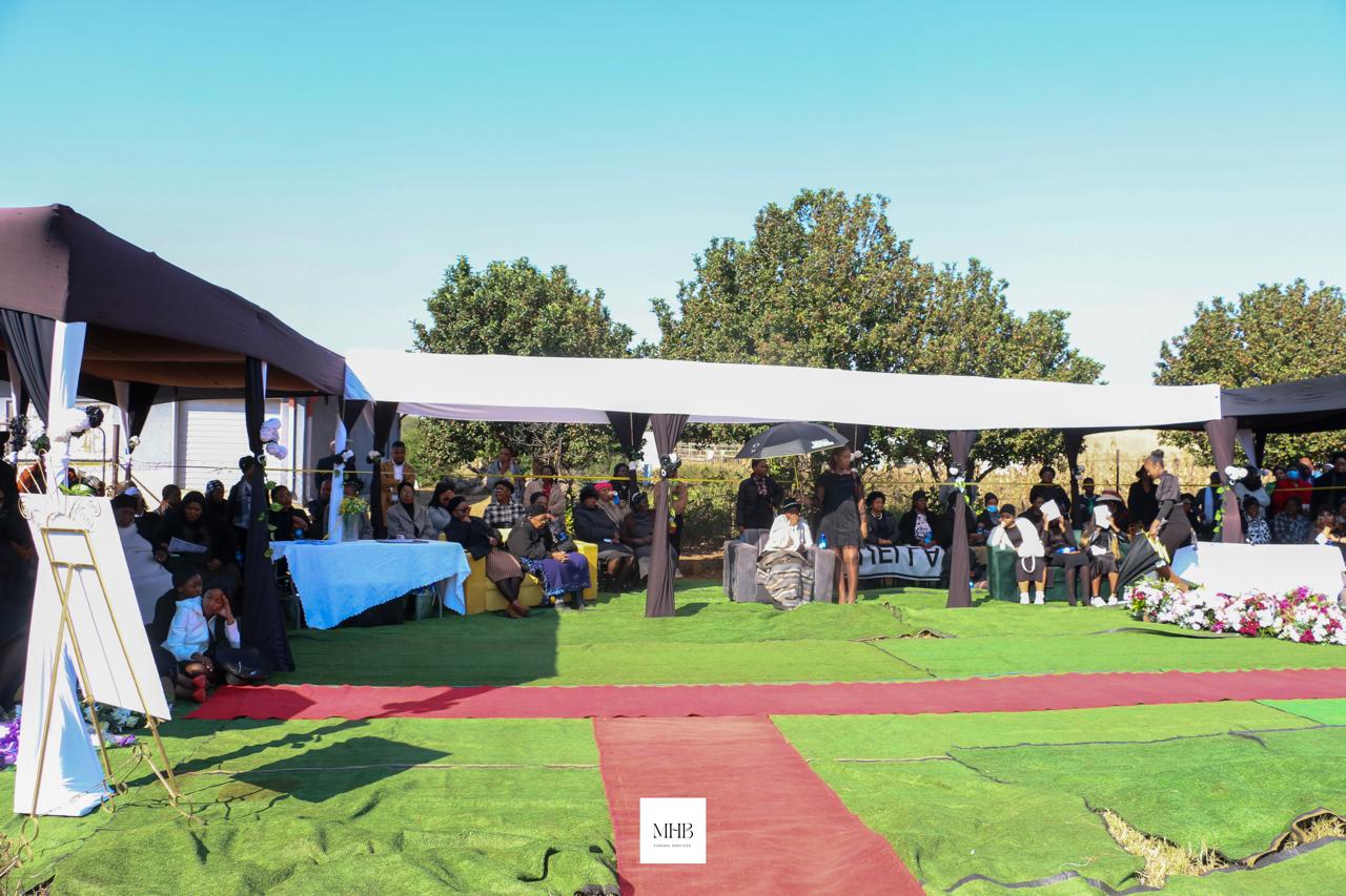 Large white and black funeral tent with seated mourners and red carpet aisle